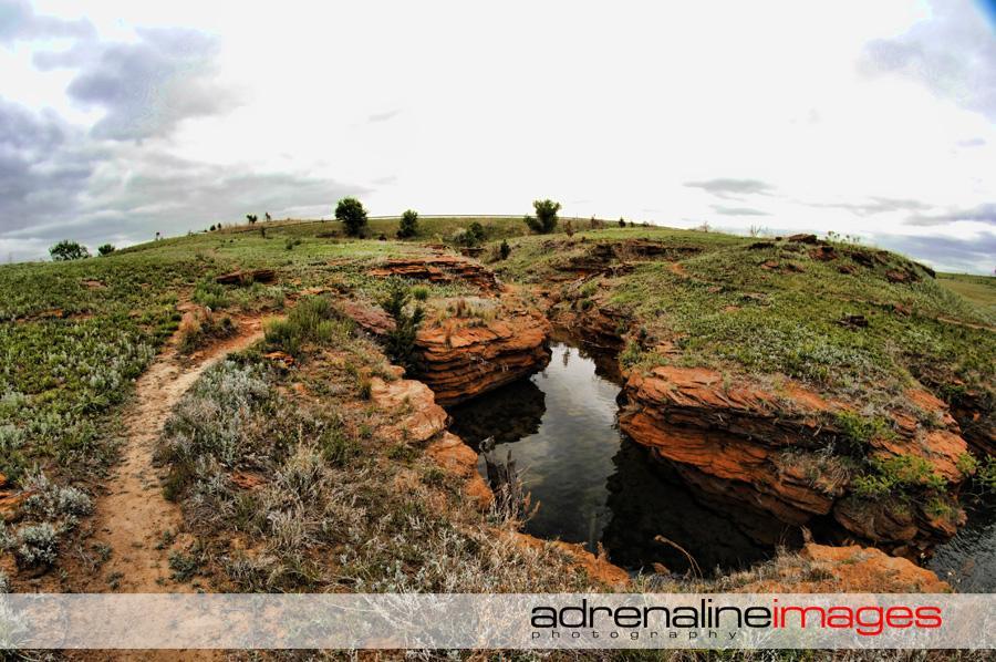 A panoramic view of a tranquil landscape featuring a winding dirt path leading to rocky terrain and a small water body surrounded by greenery. The sky is overcast, with patches of clouds, and the scene showcases undulating hills and sparse vegetation, creating a serene natural setting. Switchgrass mountain bike trail.