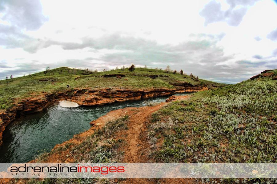 A panoramic view of a lush green landscape featuring rocky cliffs along the edge of a tranquil river. The scene includes a winding pathway leading through the grassy terrain under a cloudy sky, creating a serene natural setting. Switchgrass mountain bike trail.