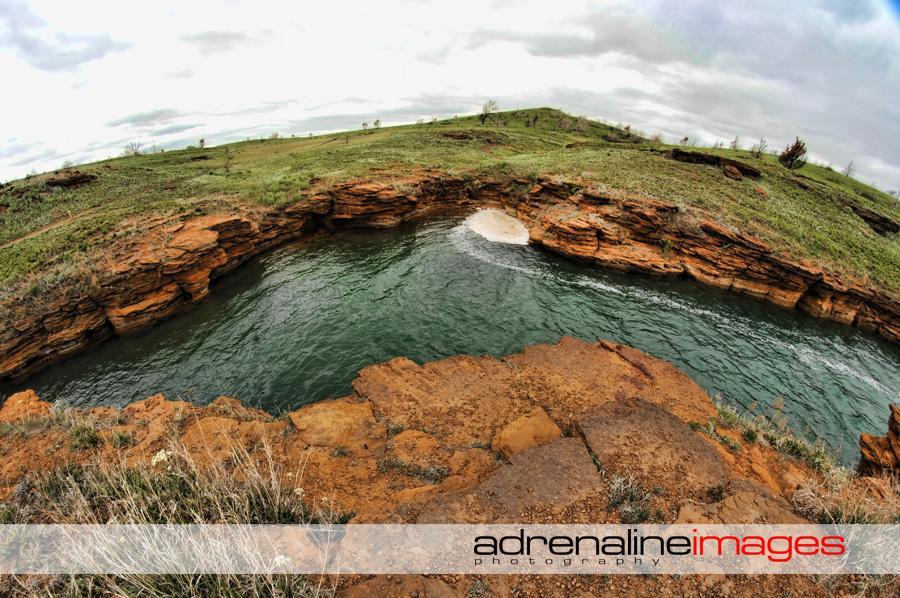A scenic view of a rocky shoreline curving around a calm body of water, surrounded by green hills under a cloudy sky. The foreground features textured rock formations, while the water reflects the overcast atmosphere, creating a tranquil natural setting. Switchgrass mountain bike trail.