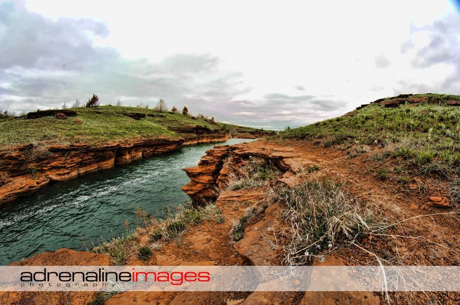 A scenic view of a river winding through rocky terrain, surrounded by grassy hills under a cloudy sky. The landscape features layers of reddish-brown rock formations alongside the water, with patches of grass and small plants visible along the riverbank. Switchgrass mountain bike trail.