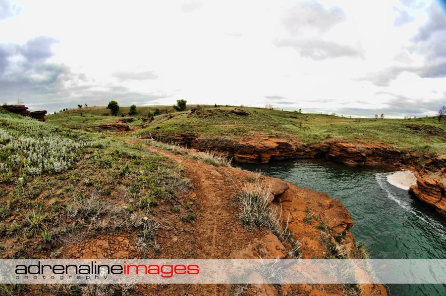 A scenic landscape featuring a rugged pathway winding along a cliffside, overlooking a calm body of water. The area is dotted with green grass and wildflowers, set against a backdrop of rolling hills and a cloudy sky. The rocky terrain showcases reddish-brown formations, emphasizing the natural beauty of the environment. Switchgrass mountain bike trail.