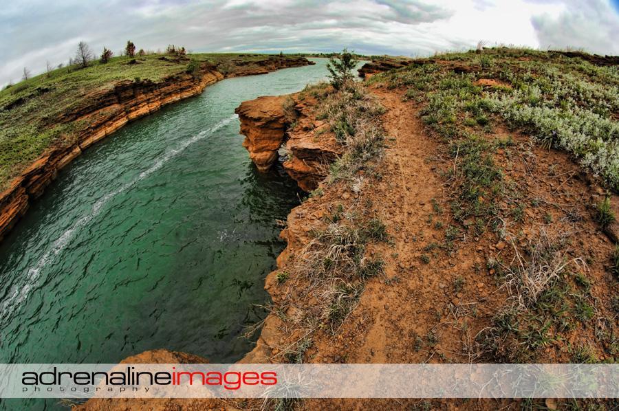 A scenic view of a winding river bordered by steep, reddish-brown cliffs, with lush green vegetation along the banks. The sky is overcast, adding a dramatic backdrop to the natural landscape. A narrow dirt path runs along the edge of the cliff, inviting exploration of the tranquil waters below. Switchgrass mountain bike trail.
