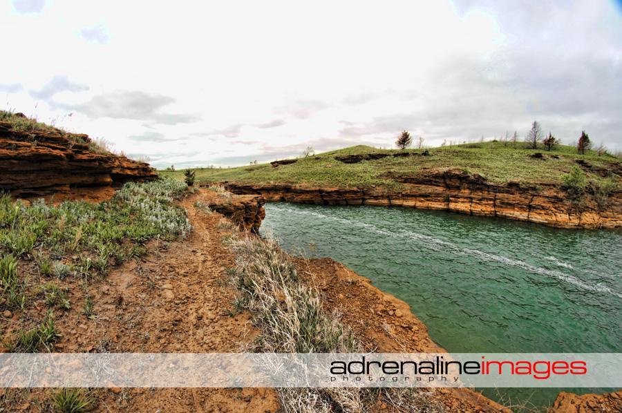 A scenic view of a river winding through a rugged terrain, featuring rocky cliffs and grassy banks under a cloudy sky. The image captures the natural beauty of the landscape, highlighting the contrasting colors of the earthy ground and the vibrant blue-green water. Switchgrass mountain bike trail.