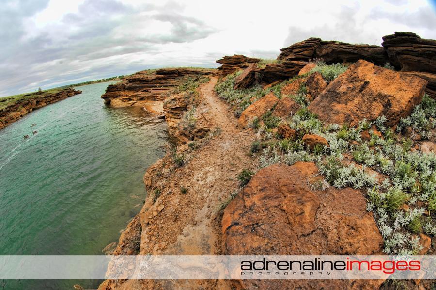 A scenic landscape featuring a winding path along a rocky shoreline with vibrant green vegetation and a calm, blue body of water. The sky is overcast, creating a dramatic atmosphere. The image captures the natural beauty of the area, highlighting the texture of the rocks and the serenity of the water. Switchgrass mountain bike trail.