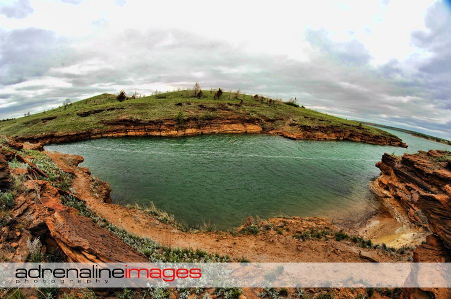 A panoramic view of a rocky shoreline surrounding a calm, greenish-blue body of water. In the background, a gently sloping green hill is dotted with sparse vegetation under an overcast sky. The landscape illustrates a natural setting with contrasting colors of the land and water. Switchgrass mountain bike trail.