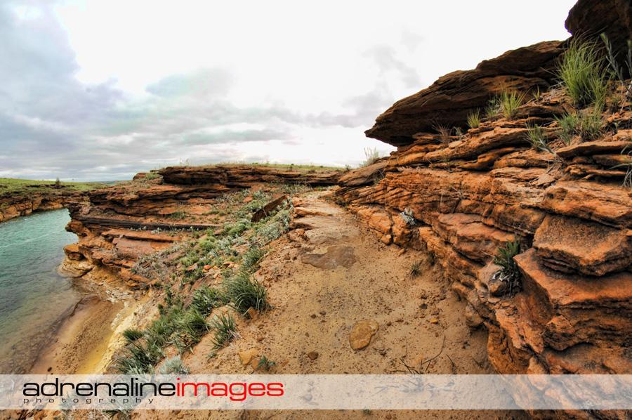A scenic landscape featuring rocky cliffs along a riverbank, with patches of greenery and sparse vegetation. The sky is overcast with gray clouds, creating a dramatic backdrop. The pathway between the rocks leads deeper into the natural setting, inviting exploration. Switchgrass mountain bike trail.
