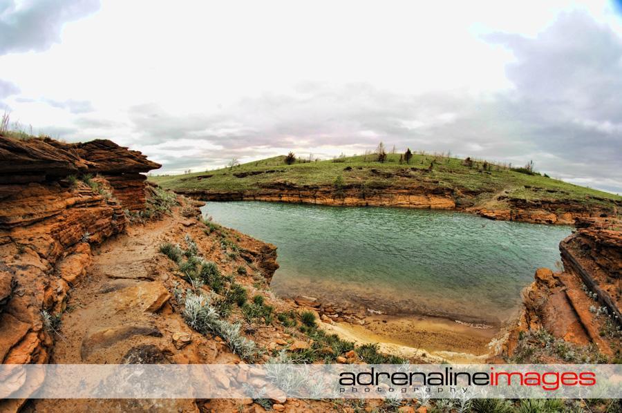 A scenic view of a river surrounded by rocky cliffs and grassy hills under a cloudy sky. The image captures the natural landscape with gentle water currents and earthy tones of the rocks and vegetation. Switchgrass mountain bike trail.
