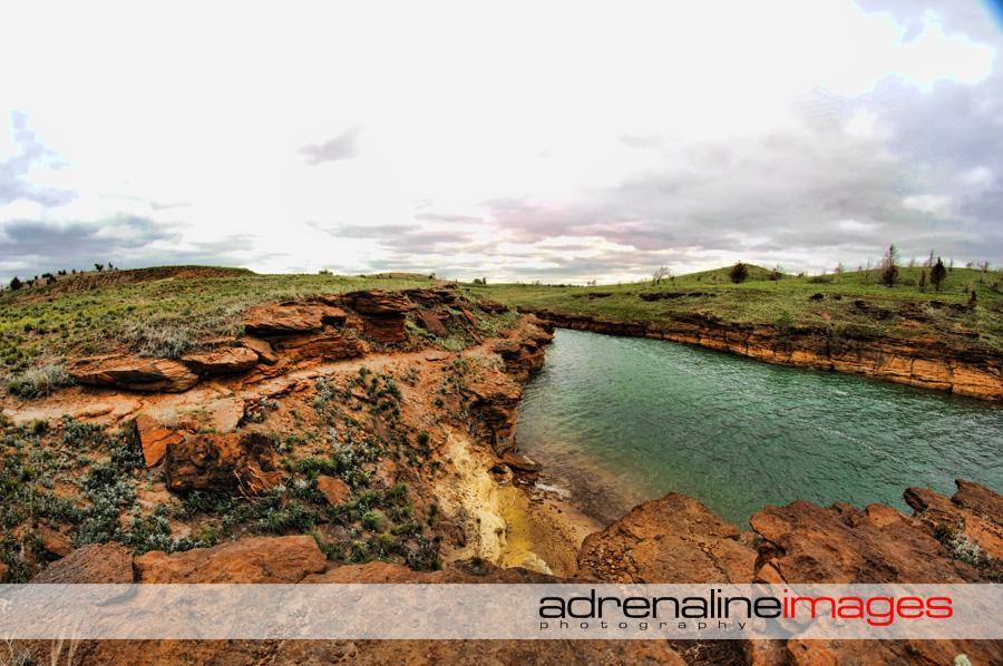 Panoramic view of a rocky shoreline alongside a calm river, with grassy hills in the background under a cloudy sky. The landscape features layers of reddish-brown rock and patches of green vegetation. Switchgrass mountain bike trail.