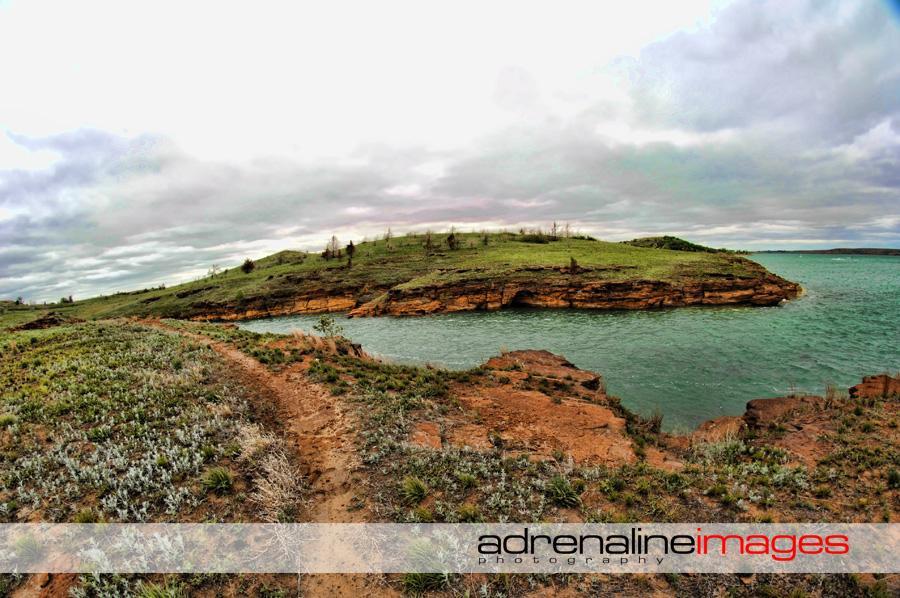 A scenic view of a rocky shoreline with a winding path leading towards a small green island in the water, under a cloudy sky. The landscape features a mix of grassy areas and rocky terrain, creating a tranquil natural setting. Switchgrass mountain bike trail.