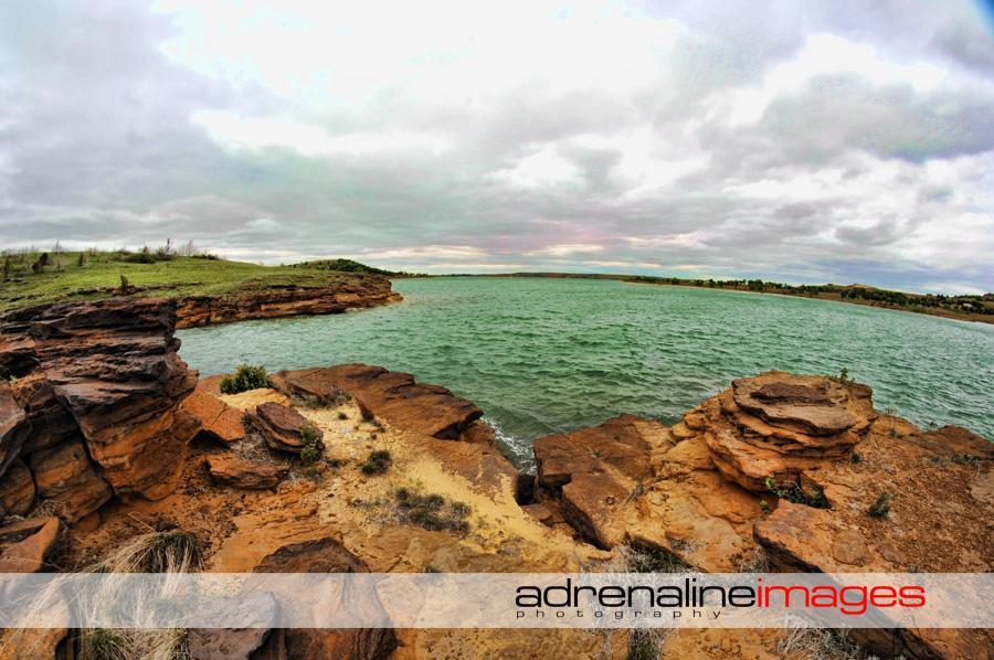 A panoramic view of a rocky shoreline with patches of grass and shrubs against a backdrop of a calm green lake. The sky is overcast with soft clouds, hinting at a serene yet slightly dramatic atmosphere. Switchgrass mountain bike trail.