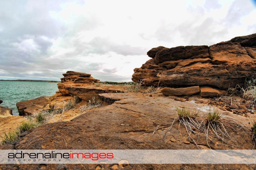 Rocky shoreline with textured brown rocks and sparse vegetation, overlooking a calm body of water under an overcast sky. The scene captures a natural landscape, highlighting the contrast between the rugged terrain and the smooth water surface. Switchgrass mountain bike trail.