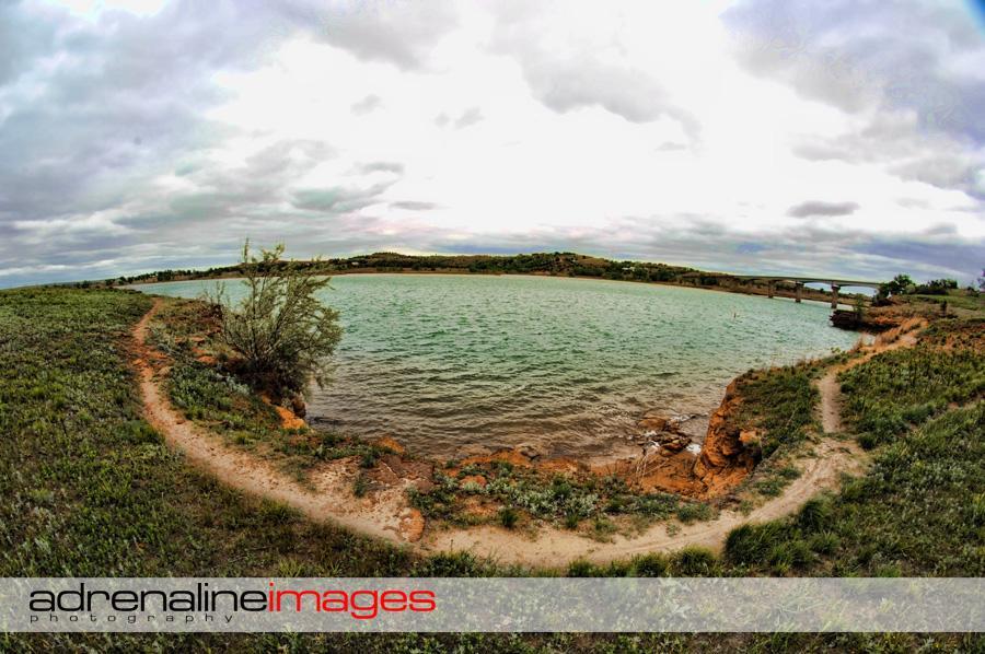 A panoramic view of a serene lakeside landscape featuring gentle green hills in the background, a turquoise body of water, and a winding dirt path along the water's edge, all under a cloudy sky. Switchgrass mountain bike trail.