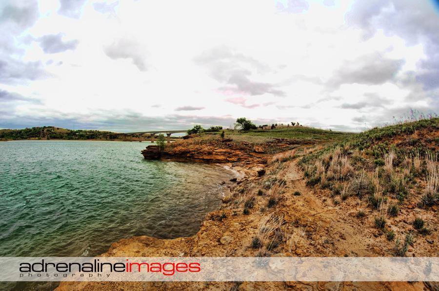 A panoramic view of a lakeshore featuring rocky terrain and lush green hills under a cloudy sky. The calm water reflects shades of blue and green, while a bridge can be seen in the background, connecting the two sides of the landscape. The foreground includes sandy soil and patches of grass, creating a natural and serene setting. Switchgrass mountain bike trail.