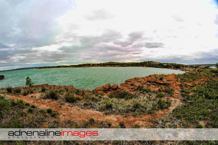A panoramic view of a calm lake surrounded by rocky cliffs and greenery under a cloudy sky, with distant hills and a bridge visible in the background. The scene captures a tranquil natural landscape. Switchgrass mountain bike trail.