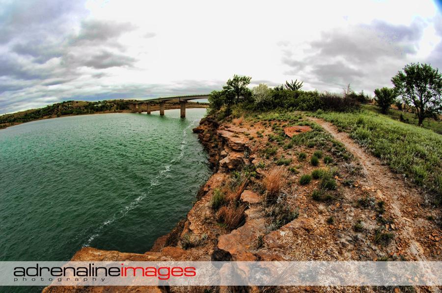 A panoramic view of a rocky shoreline next to a body of water, with a bridge spanning across in the background. The sky is overcast, and the landscape features green vegetation, shrubs, and a winding path along the rocky edge. The tranquil water reflects the surrounding scenery, creating a serene atmosphere. Switchgrass mountain bike trail.