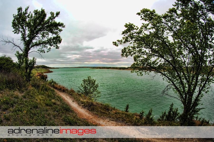 A scenic view of a calm body of water surrounded by greenery, with a winding dirt path along the shore. The sky is overcast, creating a tranquil atmosphere with shades of gray and hints of blue in the water. Tall trees frame the scene, adding to the natural beauty of the landscape. Switchgrass mountain bike trail.