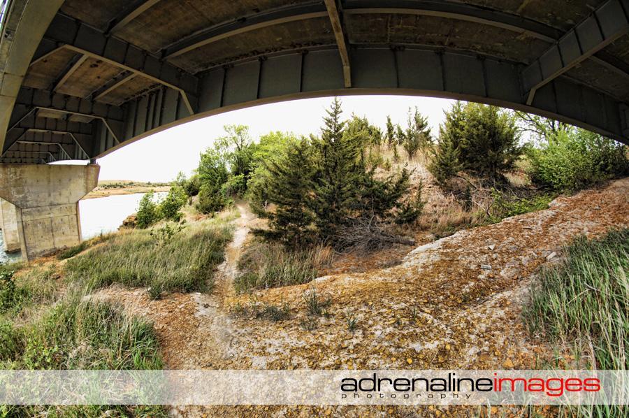 Alt text: A view from under a bridge, showing a gravel path leading towards a grassy area with small trees and shrubs. The scene includes a body of water in the background, under a cloudy sky. Switchgrass mountain bike trail.