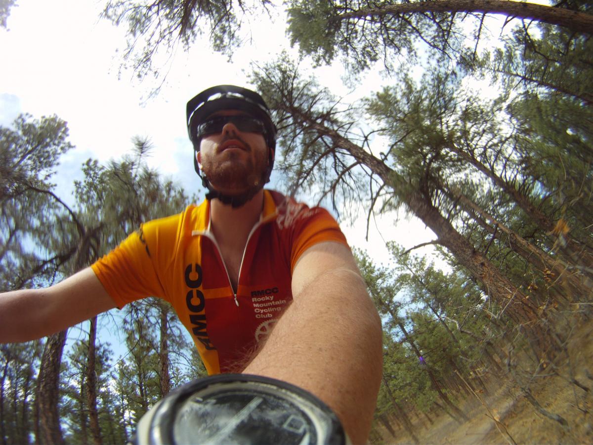 A cyclist wearing a vibrant orange and yellow jersey rides through a forested area, with tall pine trees surrounding the path. The cyclist's face is partially visible, and they are wearing a helmet and sunglasses. The handlebars and a watch can be seen in the foreground, emphasizing the action of cycling in nature. Dawson's Butte mountain bike trail.