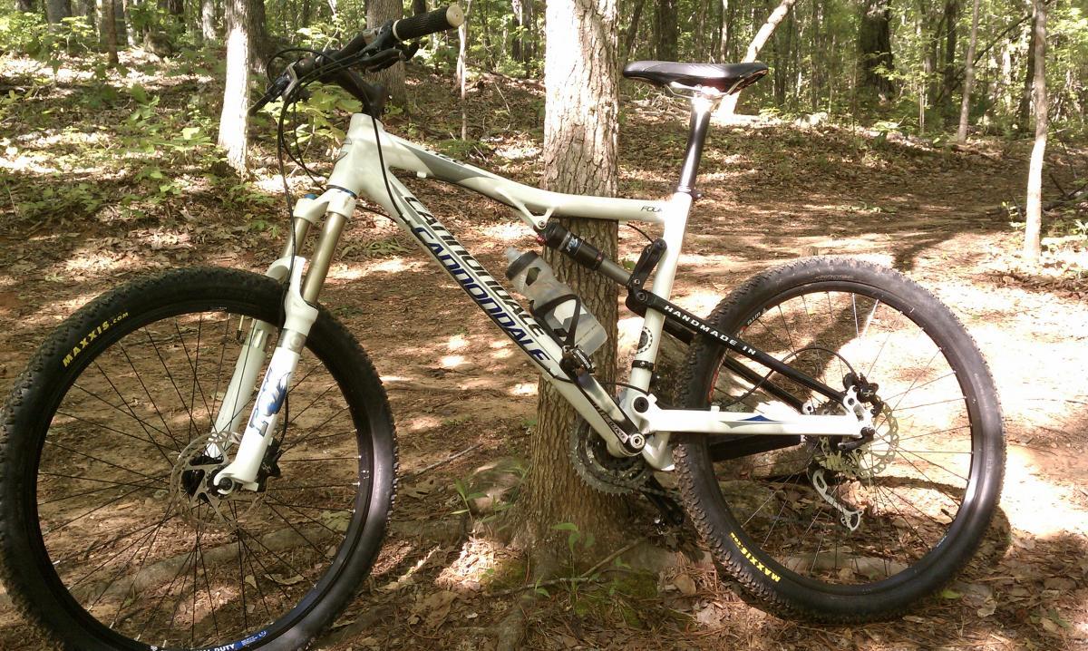 A white mountain bike leaning against a tree in a wooded area, featuring thick tires, a water bottle holder, and a visible chain and gears. The ground is covered with brown leaves and small plants, and the background shows more trees and greenery. Dauset Trails Nature Center mountain bike trail.