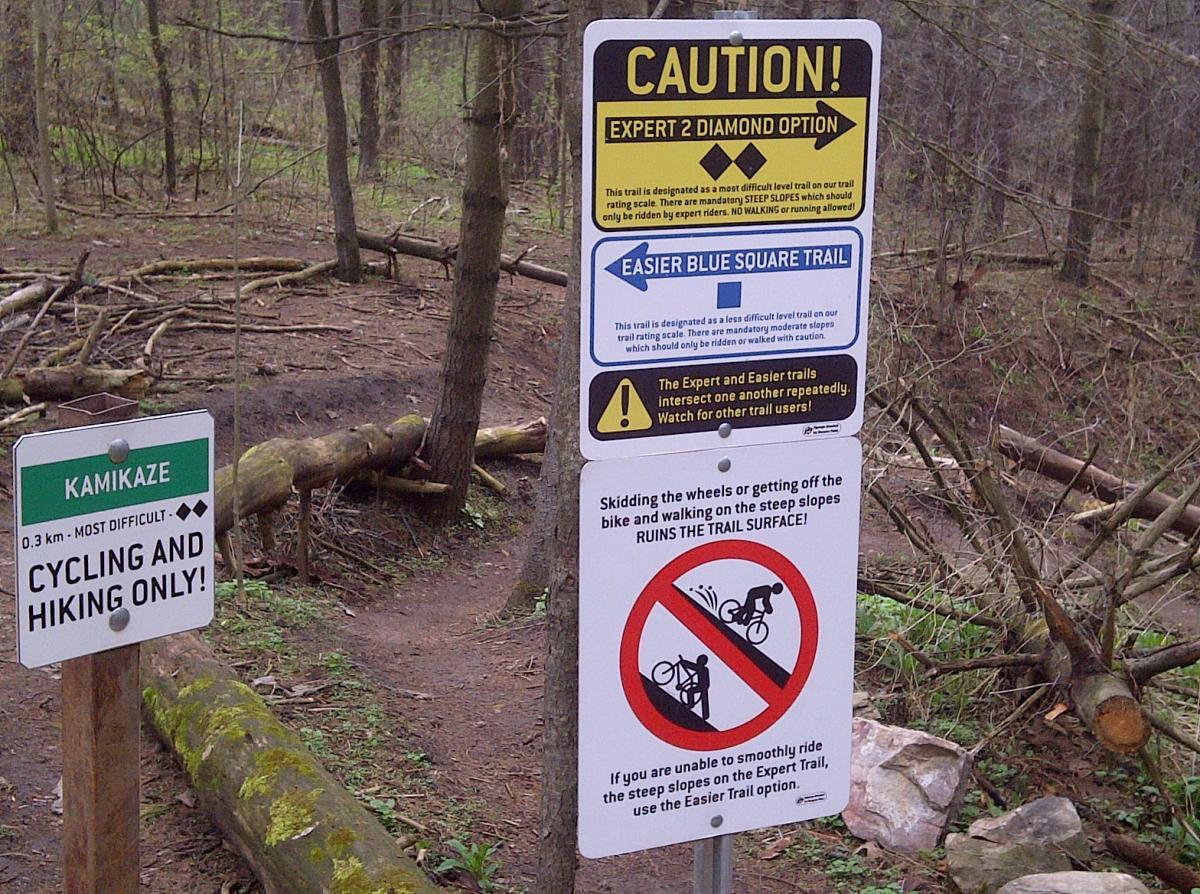 Trail signage indicating different difficulty levels for mountain biking, including an "Expert 2 Diamond Option" trail and an "Easier Blue Square Trail." The signs provide warnings about steep slopes, advise against walking or running on the Expert trail, and include a note about trail intersections. Additionally, a sign emphasizes the importance of not skidding wheels or walking on steep slopes to preserve the trail surface. Hydrocut mountain bike trail.