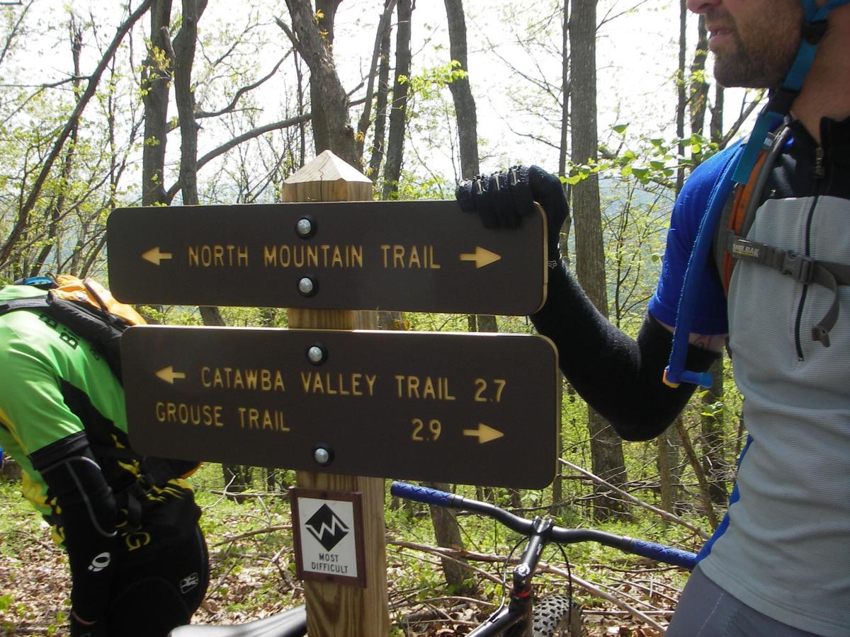 A wooden trail sign in a forest, with arrows indicating directions for three trails: North Mountain Trail, Catawba Valley Trail (2.7 miles), and Grouse Trail (2.9 miles). Two cyclists are partially visible, wearing biking gear. The surrounding area features green foliage and trees. Dragon's Back mountain bike trail.
