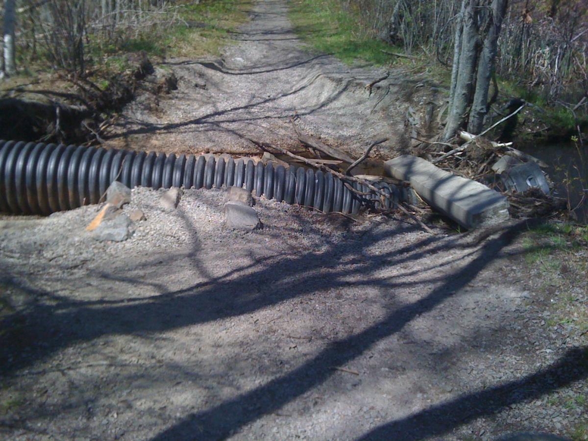 Image of a damaged path alongside a creek, featuring a large black culvert pipe obstructing the way. The ground is gravelly with scattered rocks, and the area is lined with trees casting shadows. Some debris, including branches and concrete blocks, can be seen near the pipe. Cutler Park mountain bike trail.