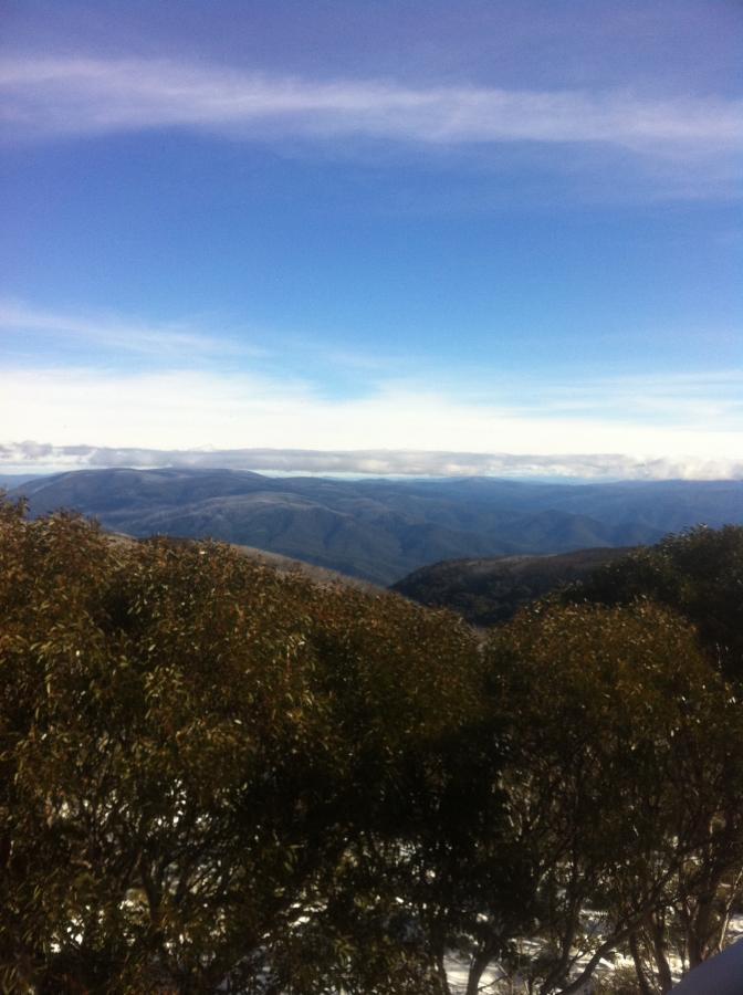 A panoramic view of rolling mountains under a clear blue sky, with a few white clouds. In the foreground, there are green trees partially obscuring the view of the landscape beyond. The scenery suggests a natural, serene environment typical of a mountainous region. Cascade Hut Trail mountain bike trail.