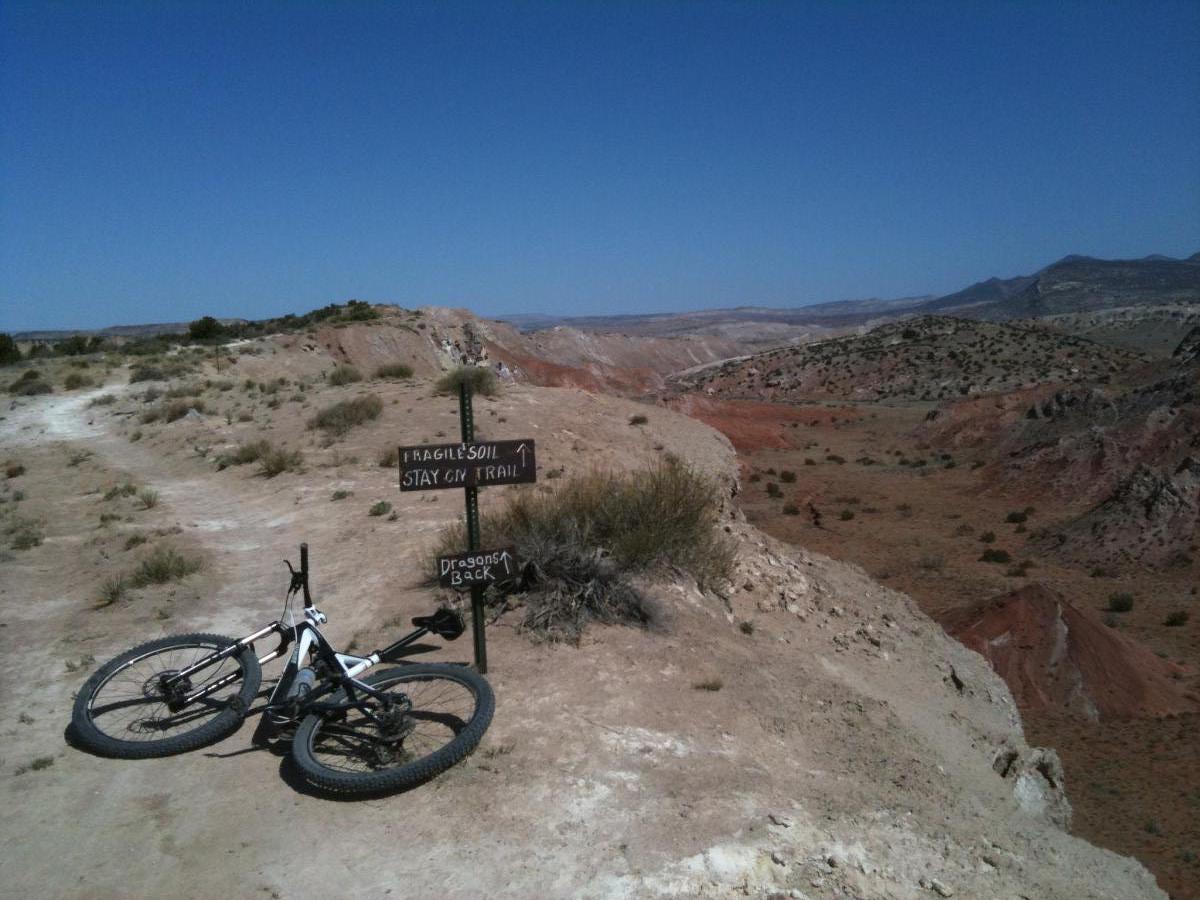 A mountain bike rests on a dirt trail near a warning sign that reads "Fragile Soil - Stay on Trail" and points toward "Dragon's Back." The landscape features rugged, red and brown hills under a clear blue sky, highlighting the natural beauty of the area. White Ridge Bike Trails mountain bike trail.