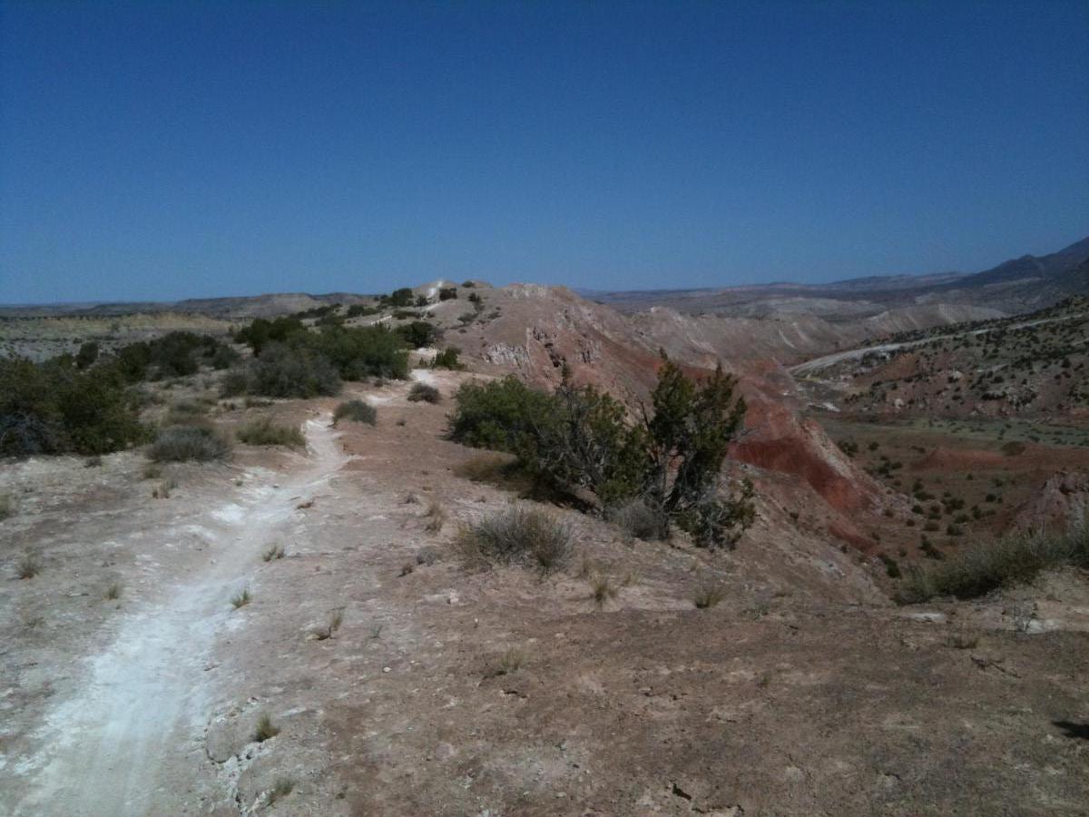 A panoramic view of a dry, rugged landscape featuring hills and mesas under a clear blue sky. A winding dirt path leads through sparse vegetation and rocky terrain, with distant mountains visible on the horizon. White Ridge Bike Trails mountain bike trail.