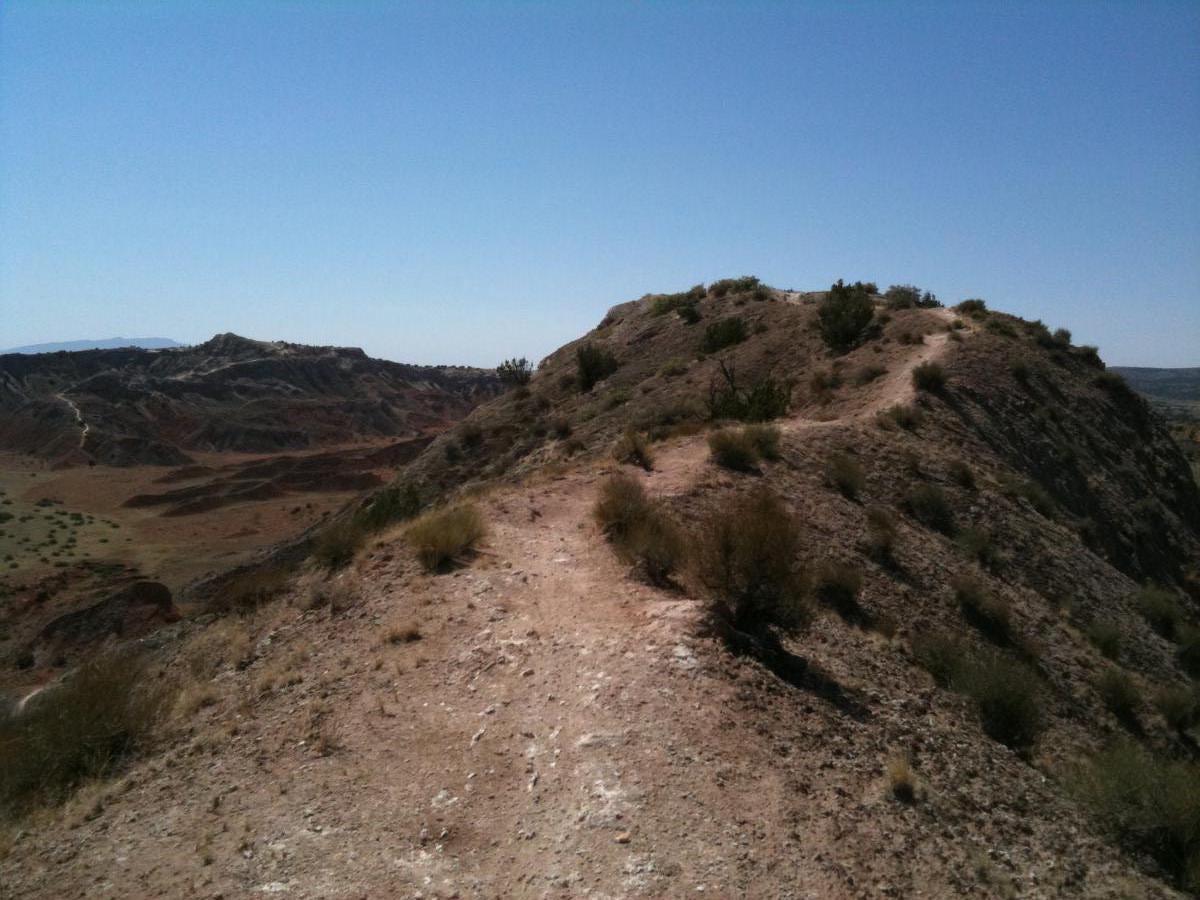 A rocky landscape featuring a winding dirt path leading up a hill, surrounded by rugged terrain and sparse vegetation under a clear blue sky. White Ridge Bike Trails mountain bike trail.