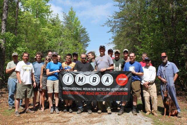 A group of people standing together in a forested area, holding a banner for the International Mountain Bicycling Association (IMBA) and the Southern Off-Road Bicycle Association (SORBA). The individuals are dressed casually, suitable for outdoor activities, and the scene is sunny with greenery in the background. A dog is also present among the group. Paynes Creek mountain bike trail.