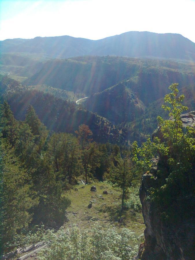 A scenic view of a mountainous landscape, featuring rolling hills and lush greenery under a bright sky. Sunlight filters through the trees, illuminating the valley below, which includes a winding road and a diverse array of vegetation. The scene conveys a sense of tranquility and natural beauty. Jardine Juniper mountain bike trail.