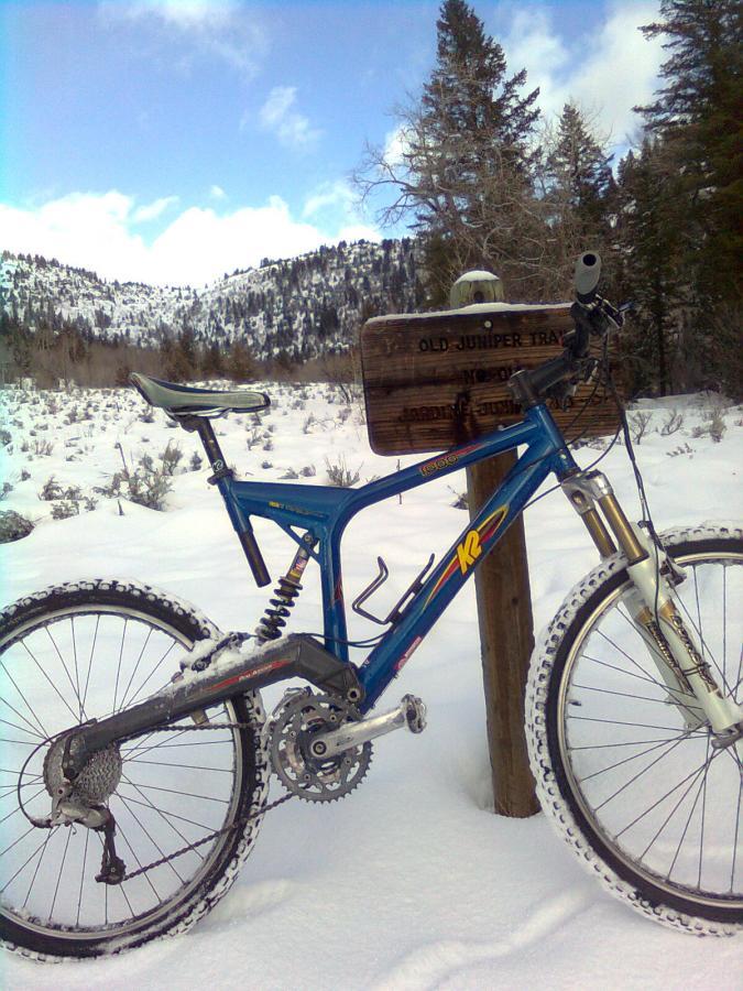 A blue mountain bike covered in snow is parked next to a wooden trail sign labeled "Old Juniper Trail." The background features snow-covered mountains and trees under a partly cloudy sky. Jardine Juniper mountain bike trail.