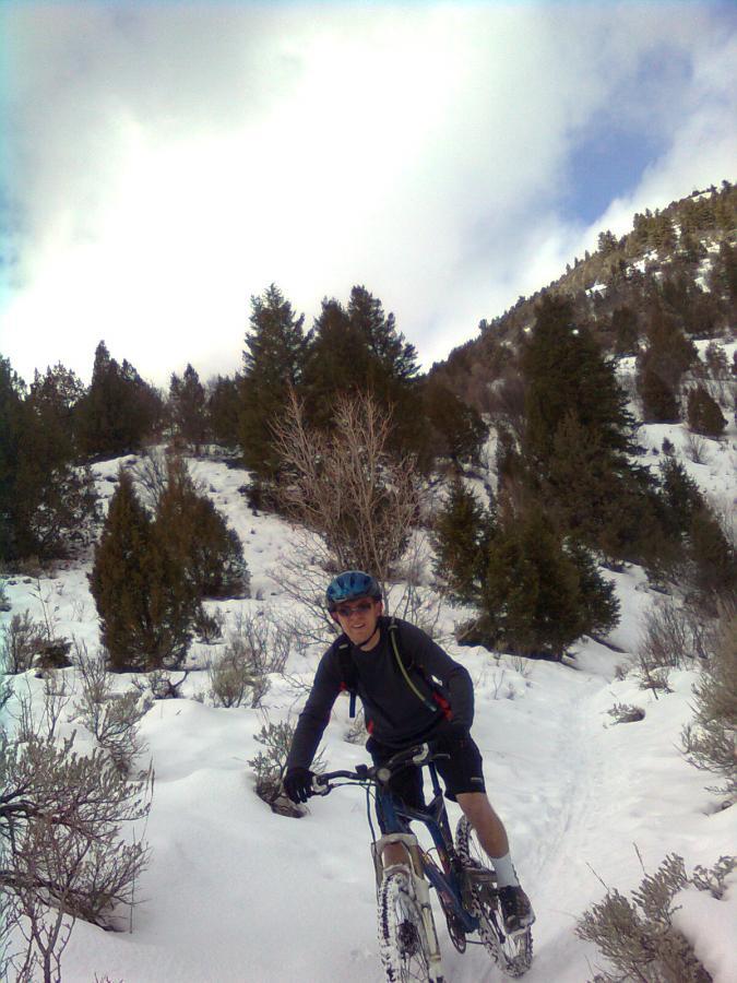 A person riding a mountain bike on a snowy trail surrounded by trees and hills, wearing a blue helmet and athletic clothing. The sky is partly cloudy, creating a bright, outdoor atmosphere. Jardine Juniper mountain bike trail.