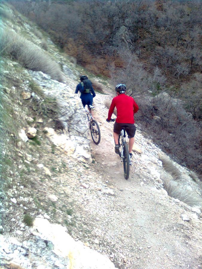 Two mountain bikers ride along a narrow, rocky trail surrounded by sparse vegetation and a steep drop-off on one side. One cyclist is wearing a red shirt and shorts, while the other is dressed in a dark top with a backpack. The terrain appears rugged, indicative of a challenging biking path. Bonneville Shoreline Trail - Ogden Section mountain bike trail.