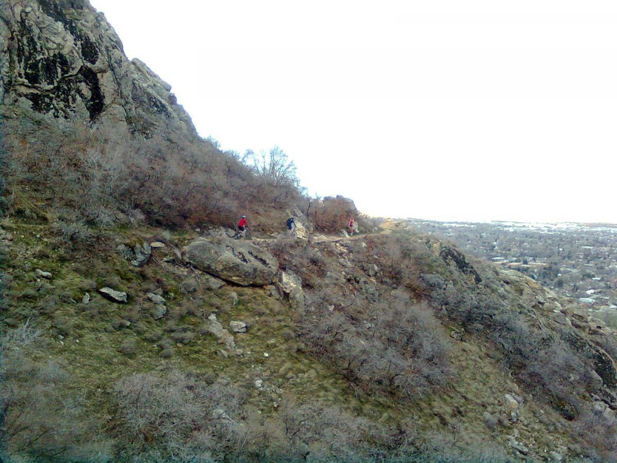 A rocky hillside with patches of grass, featuring several hikers making their way along a narrow trail. The background shows a distant view of a town below, with a mix of trees and shrubs scattered across the landscape. The sky is overcast, creating a muted atmosphere. Bonneville Shoreline Trail - Ogden Section mountain bike trail.