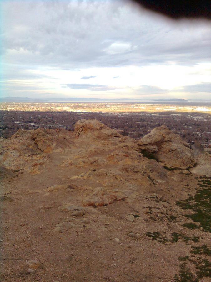 A rocky outcrop overlooking a vast valley, with scattered greenery and a cloudy sky above. The view includes distant hills and a cityscape in the background, suggesting a mountainous region. Bonneville Shoreline Trail - Ogden Section mountain bike trail.