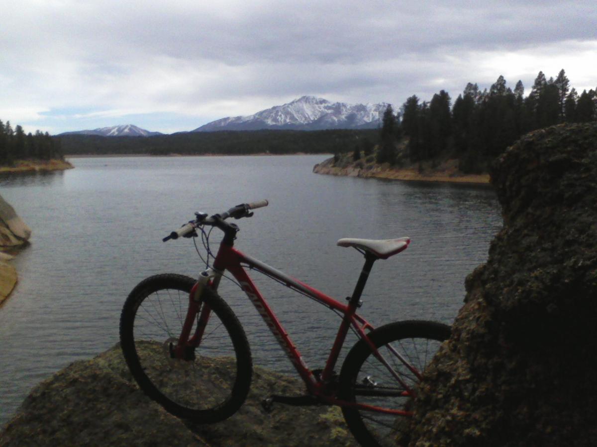 A red mountain bike is leaning against a large rock at the edge of a serene lake, with snow-capped mountains and pine trees visible in the background under a cloudy sky. Rampart Reservoir mountain bike trail.
