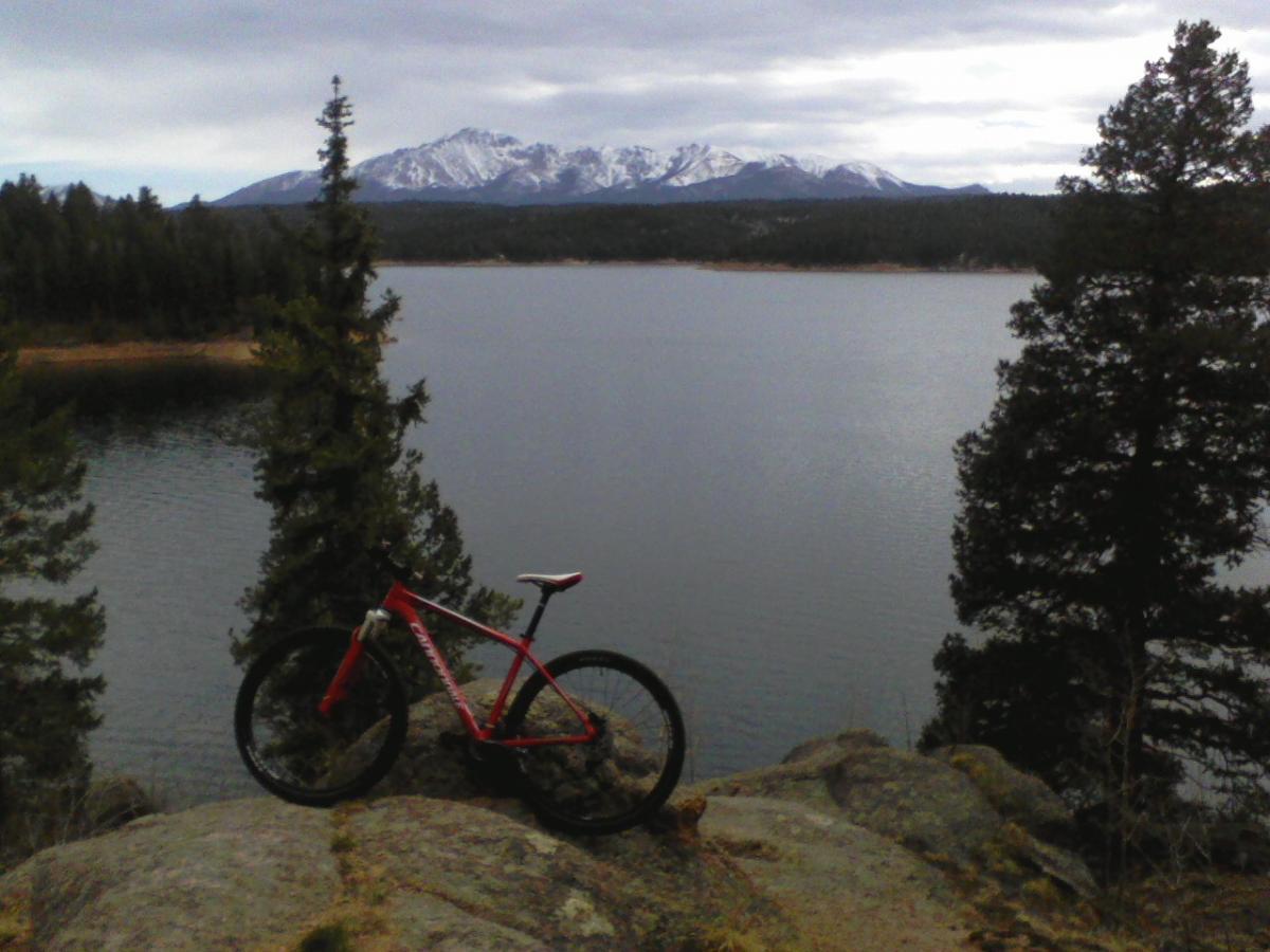 A red mountain bike rests on a rocky outcrop overlooking a calm lake, with a backdrop of snow-capped mountains and a forested shoreline under a cloudy sky. Rampart Reservoir mountain bike trail.