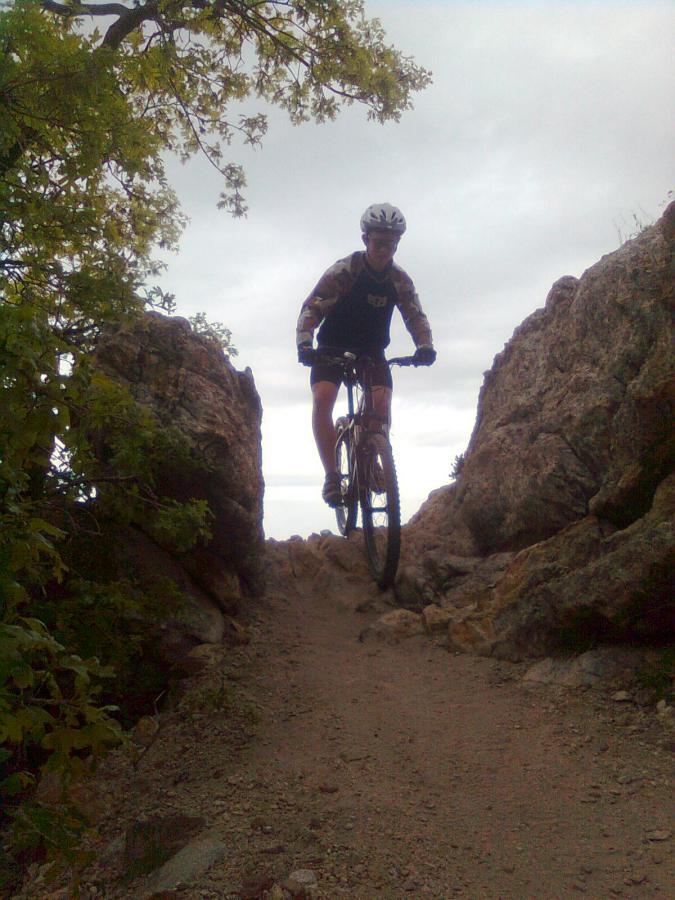 A mountain biker navigating a rocky trail between two large boulders, with greenery and a cloudy sky in the background. Bonneville Shoreline Trail - Ogden Section mountain bike trail.