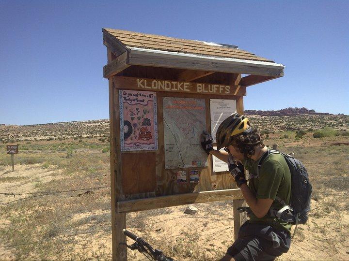 A mountain biker in a helmet studies a trail map at the Klondike Bluffs trailhead, surrounded by arid terrain under a clear blue sky. A wooden information board displays various maps and notices, with desert landscape visible in the background. Klondike Bluffs mountain bike trail.