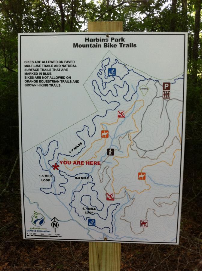 A detailed trail map for Harbins Park, featuring mountain bike trails designated with blue lines and notes indicating distances. The map highlights areas where biking is allowed and where it is prohibited, marked by corresponding symbols. The location "YOU ARE HERE" is clearly indicated in red. Surrounding details include a north arrow and symbols for various recreational activities. Harbins Park mountain bike trail.