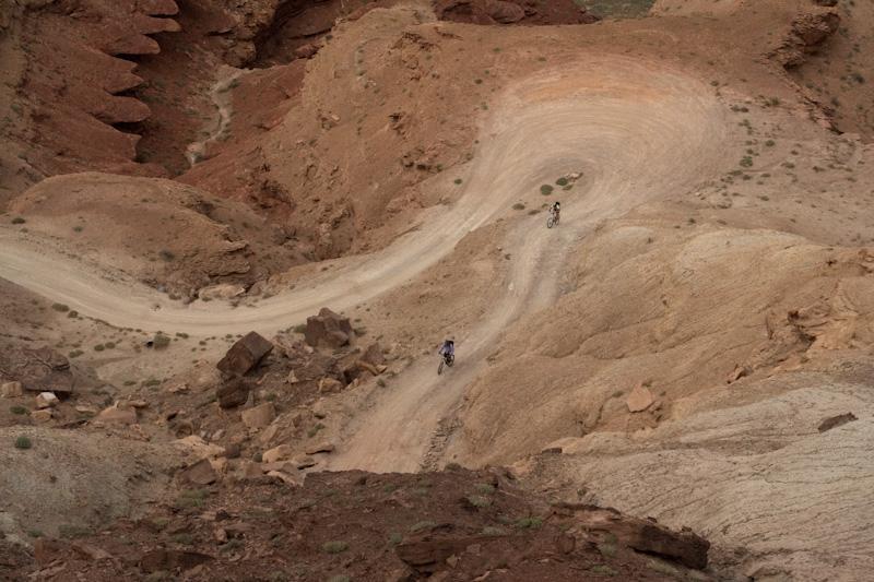 Two mountain bikers navigate a winding dirt trail through a rugged, rocky landscape with various shades of brown and orange. Sparse vegetation is visible among the rocky terrain, highlighting the natural beauty of the area. White Rim Trail mountain bike trail.