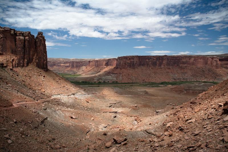 A panoramic view of a vast canyon landscape featuring towering reddish cliffs under a partly cloudy blue sky. The lower area displays rocky terrain and sparse vegetation, with a winding path visible in the foreground leading towards the canyon bottom. White Rim Trail mountain bike trail.