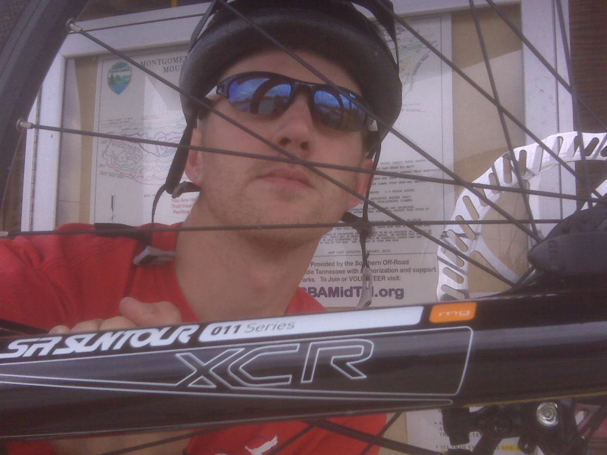 A person wearing sunglasses and a helmet holds a mountain bike, with the bike's front wheel and spokes visible in the foreground. In the background, there is a map and informational poster about Montgomery Bell State Park. The individual is wearing a red shirt and appears to be looking at the camera. Montgomery Bell State Park Mtb Trail mountain bike trail.