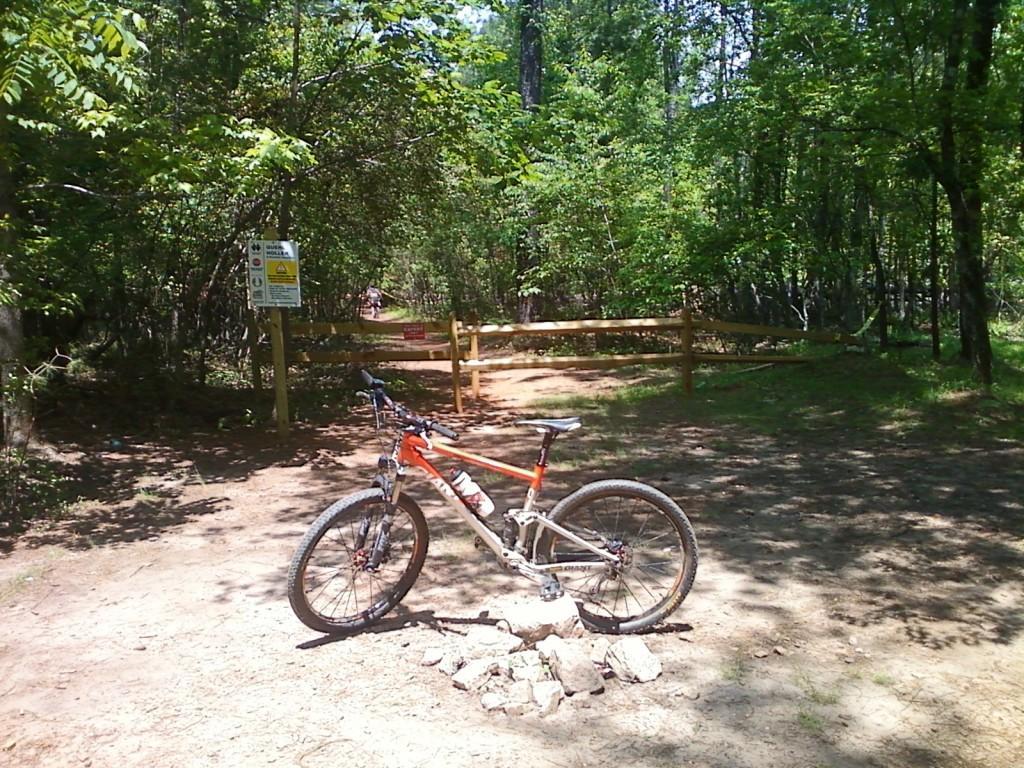 A mountain bike resting on a rock formation at a trailhead in a wooded area. The path leads into a forest with lush green trees, and a sign indicating trail rules and warnings is visible near a wooden fence in the background. Quehl Holler mountain bike trail.