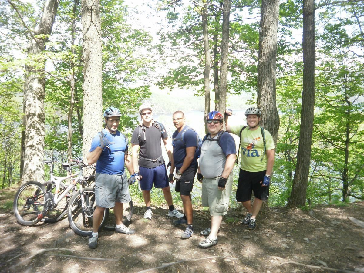 A group of five men stand together on a forested trail, with bicycles leaning against trees beside them. They are dressed in casual cycling attire, smiling at the camera. The background features lush green trees and a glimpse of water through the foliage, indicating a scenic outdoor setting. Allegrippis Trails mountain bike trail.