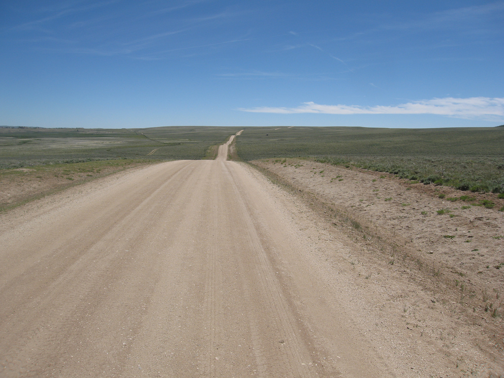 Dirt road stretching through an open landscape under a clear blue sky, surrounded by sparse vegetation and rolling hills in the background.