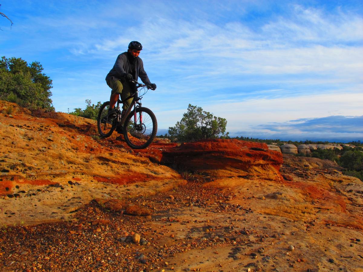 A mountain biker navigating a rocky trail on a sunny day, surrounded by natural scenery and a blue sky with wispy clouds. The terrain features reddish-orange rock formations and sparse vegetation in the background. Gooseberry Mesa mountain bike trail.