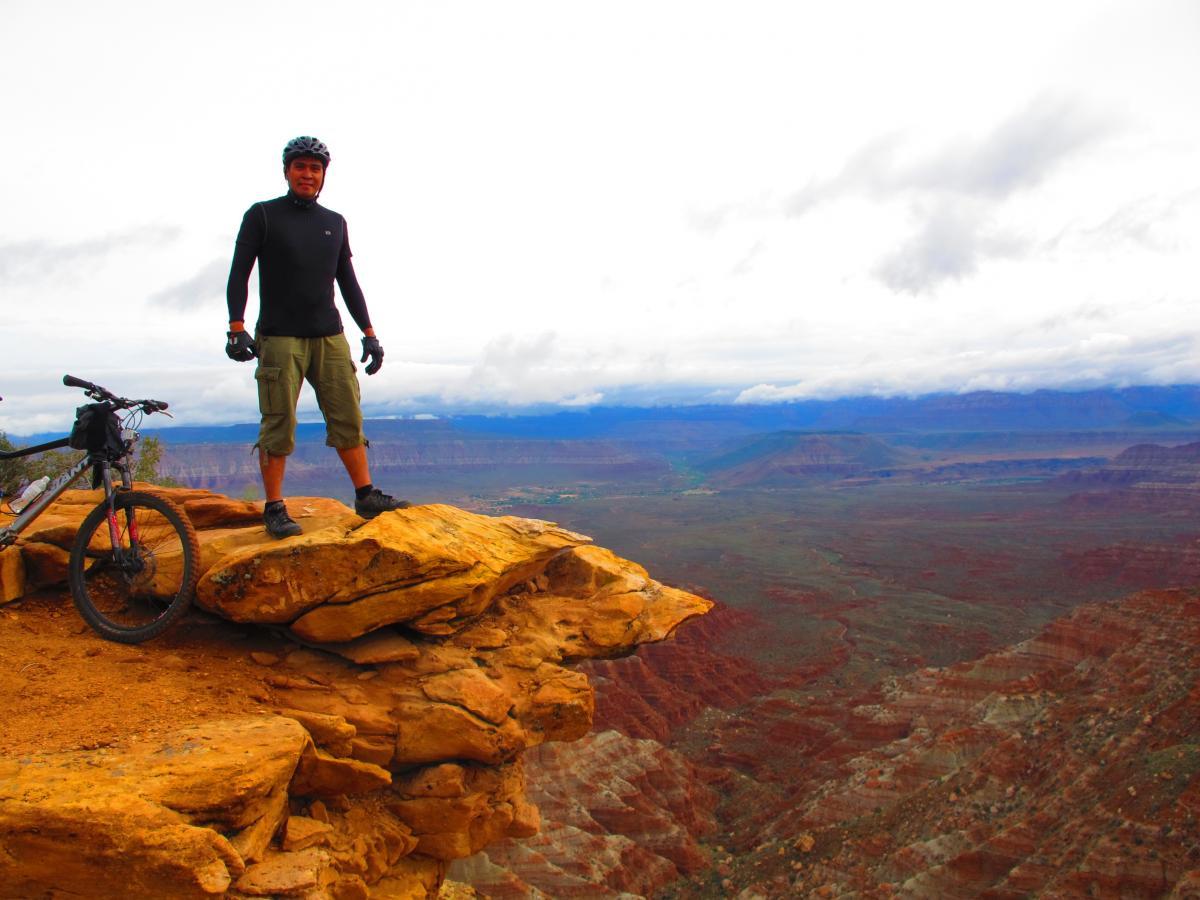 A mountain biker standing confidently on a rocky cliff edge, with a mountain bike resting beside him. The expansive landscape features layers of red and brown rock formations under a cloudy sky. Gooseberry Mesa mountain bike trail.